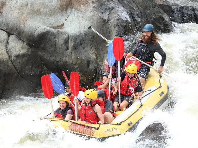A group of people white water rafting through rapids on a yellow raft.